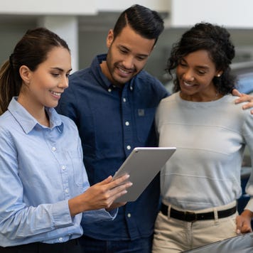 Saleswoman in blue shirt at a car dealership holds a tablet while talking to a smiling couple.