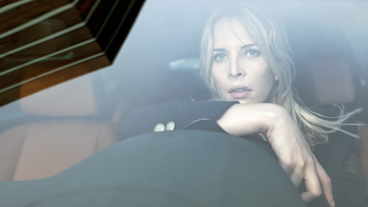 Blonde woman stares out windshield while leaning on steering wheel.