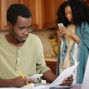 Man sits at kitchen table with pen and paper while woman stand behind on cell phone