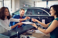 Woman to the left accepting pen and clipboard from other woman across desk, man sitting to the left as well, all smiling in dealership