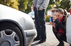 A woman taking a picture of the front of her dented car with her smartphone after an accident.