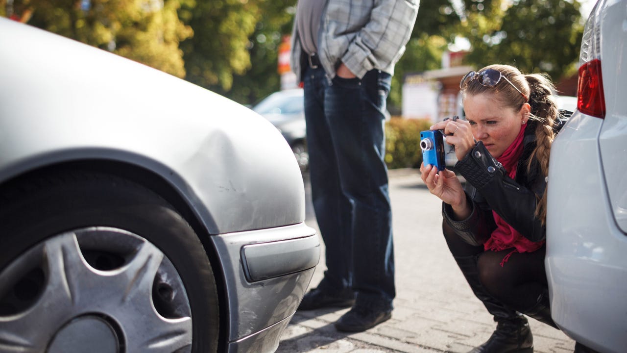 A woman taking a picture of the front of her dented car with her smartphone after an accident.