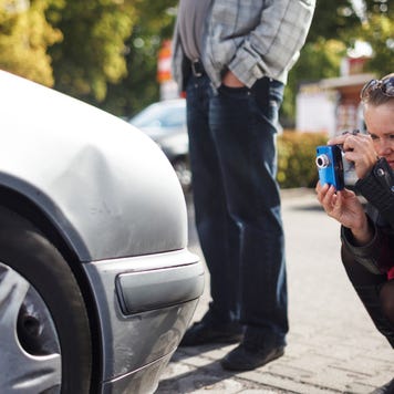A woman taking a picture of the front of her dented car with her smartphone after an accident.