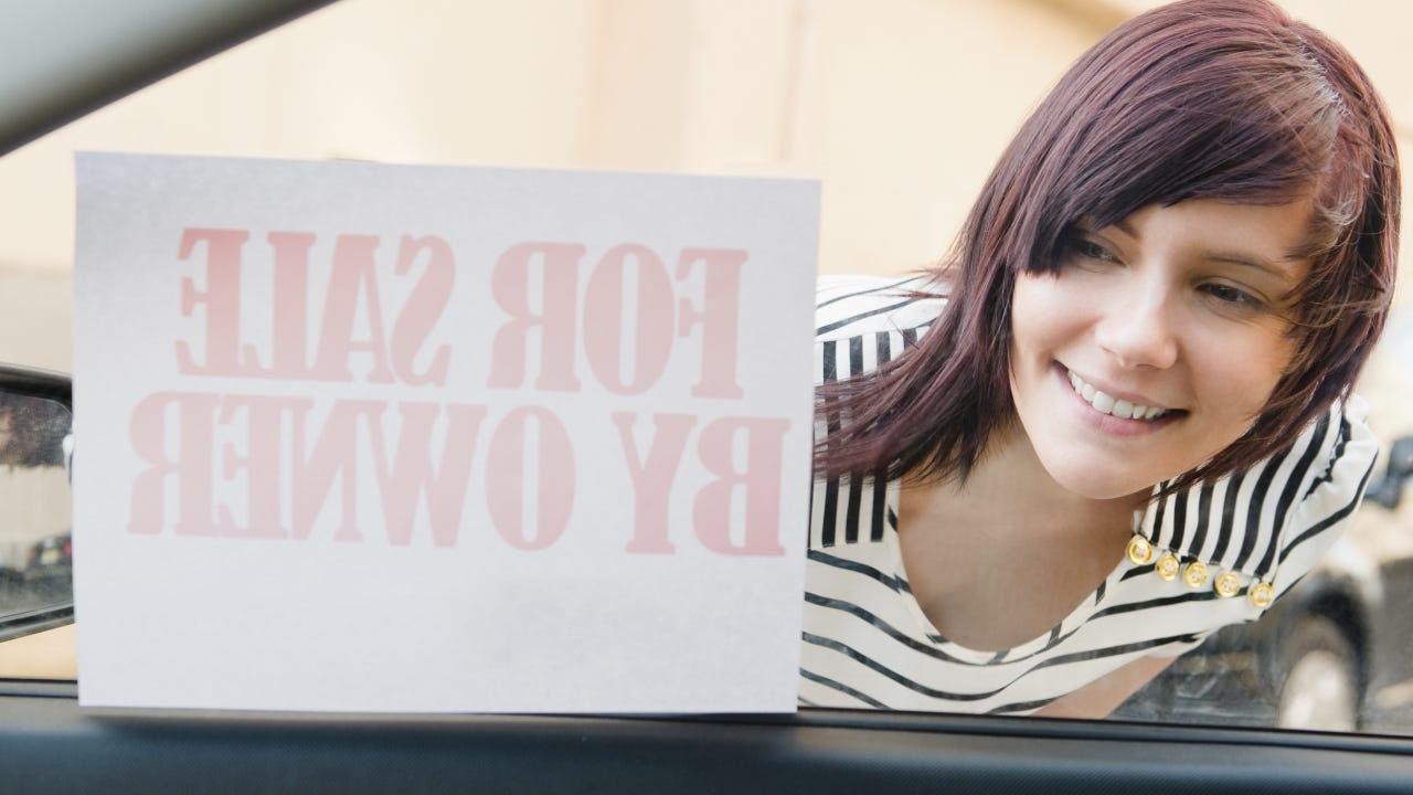 Young woman looking in car window, sign that reads for sale by owner to the left of woman