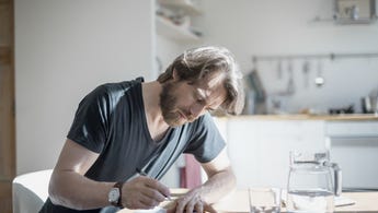 Portrait of bearded man sitting at a table at home filling out a document with pitcher and glass on the table in front of him