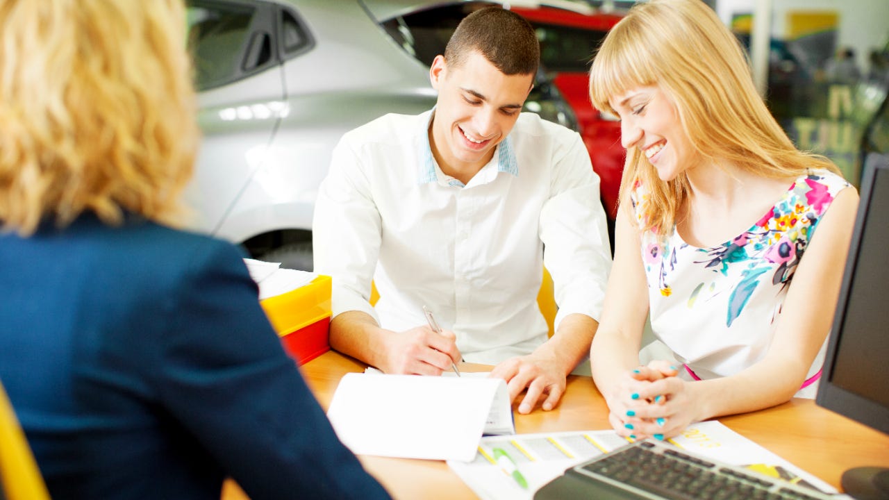 Man and woman sitting at desk in car dealership, smiling while man signs documents