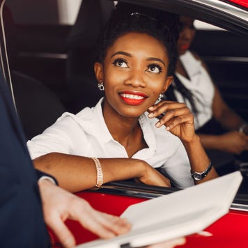 Two Black women sit in a red car at a dealership. There is a third person out of the frame holding documents.
