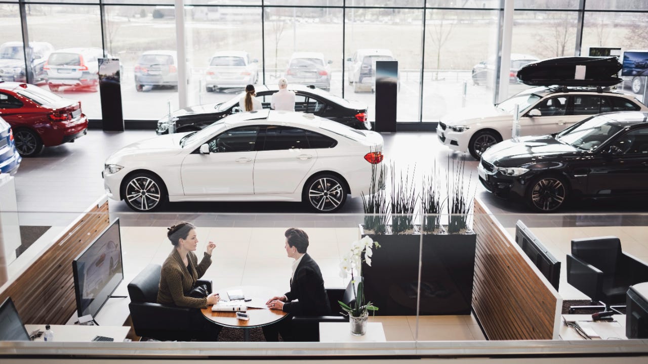 Wide shot of a car showroom with two people looking at a car in the background and two reviewing documents at a desk in the foreground