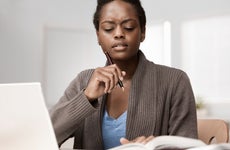 Woman with pen frowns at open book and laptop
