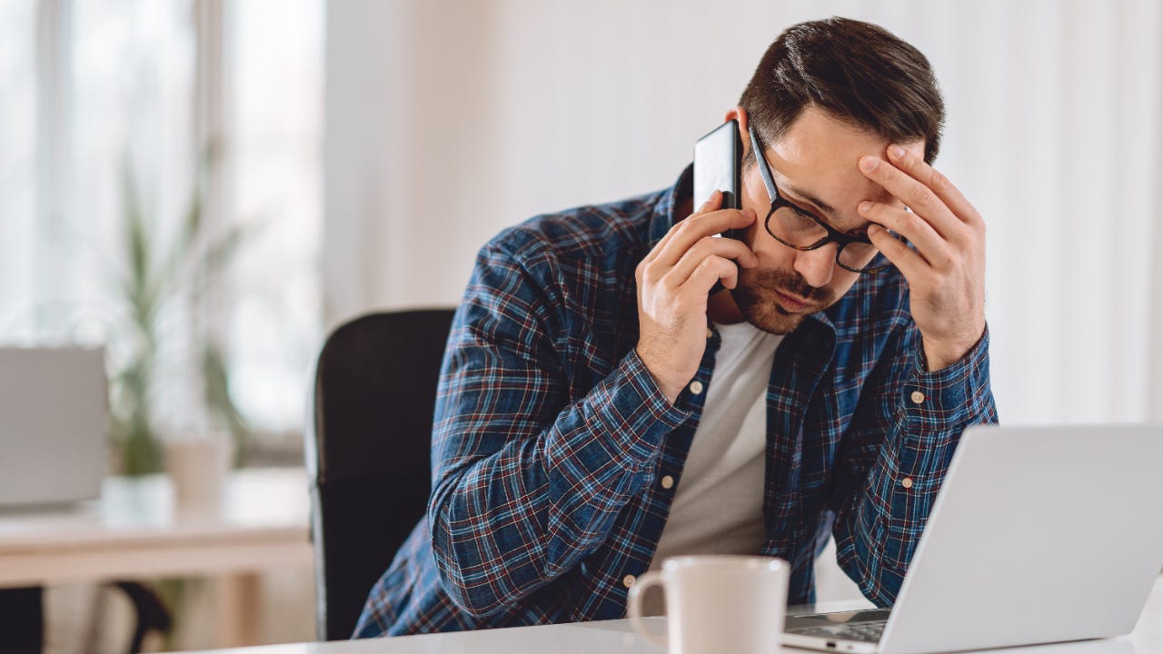 Upset looking man in glasses on phone in front of laptop with coffee mug nearby