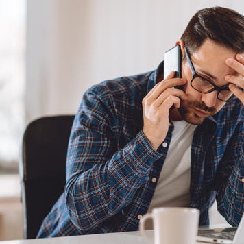 Upset looking man in glasses on phone in front of laptop with coffee mug nearby