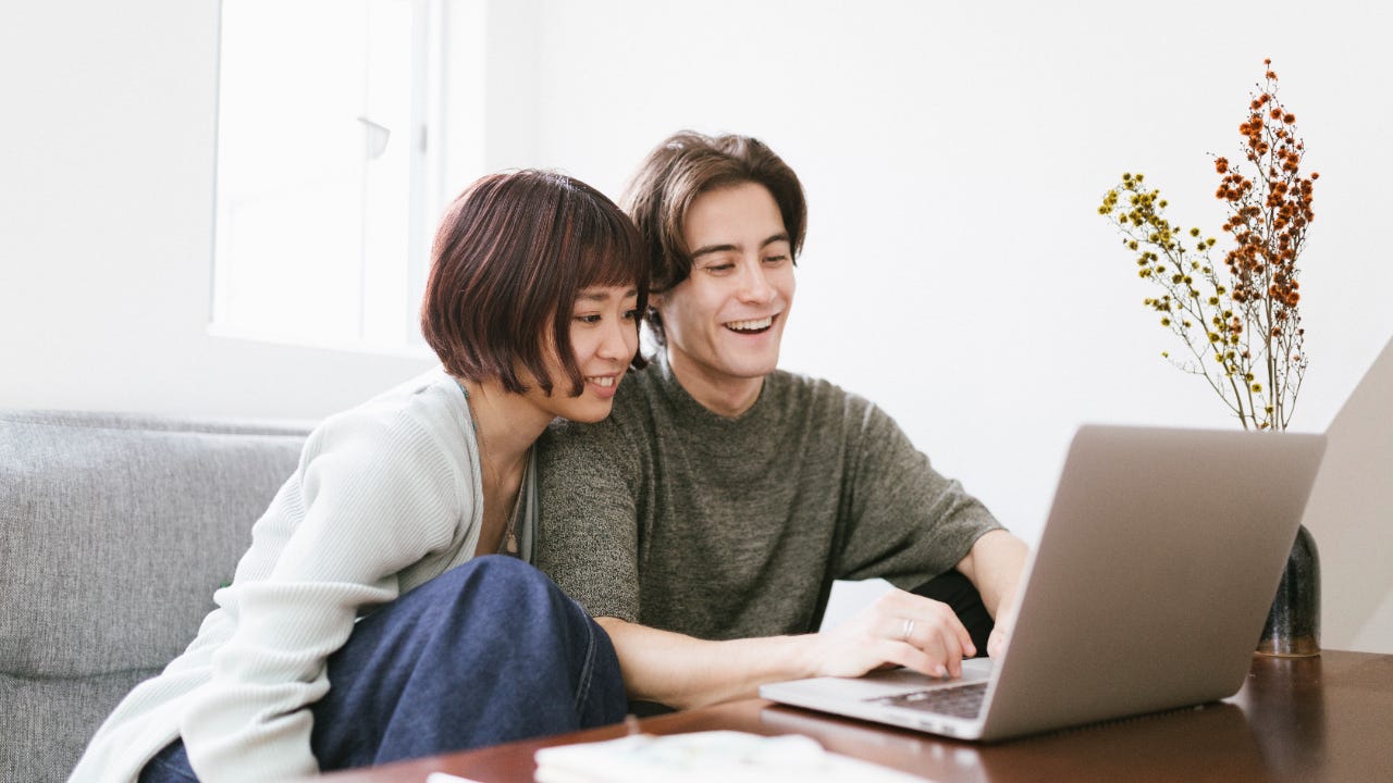 Woman leaning over man's shoulder, both smiling and looking at a laptop