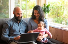 A husband and wife sit on the couch with their infant child in front of a laptop and try to figure out how to save.