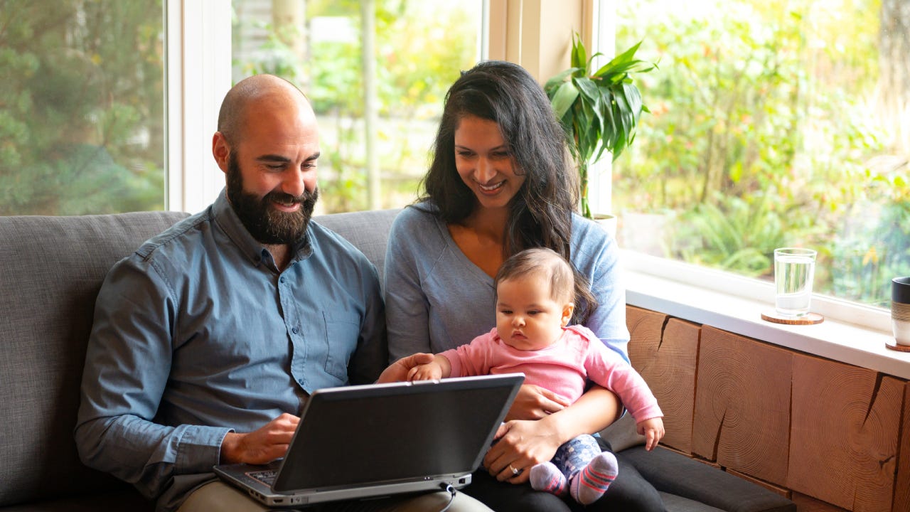 A husband and wife sit on the couch with their infant child in front of a laptop and try to figure out how to save.