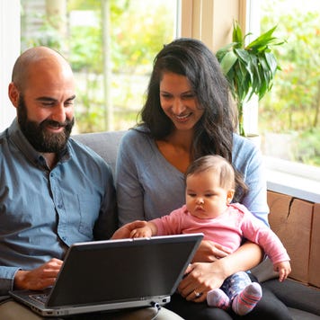 A husband and wife sit on the couch with their infant child in front of a laptop and try to figure out how to save.