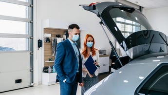 Man and woman standing in a garage with masks on looking in the trunk of a car