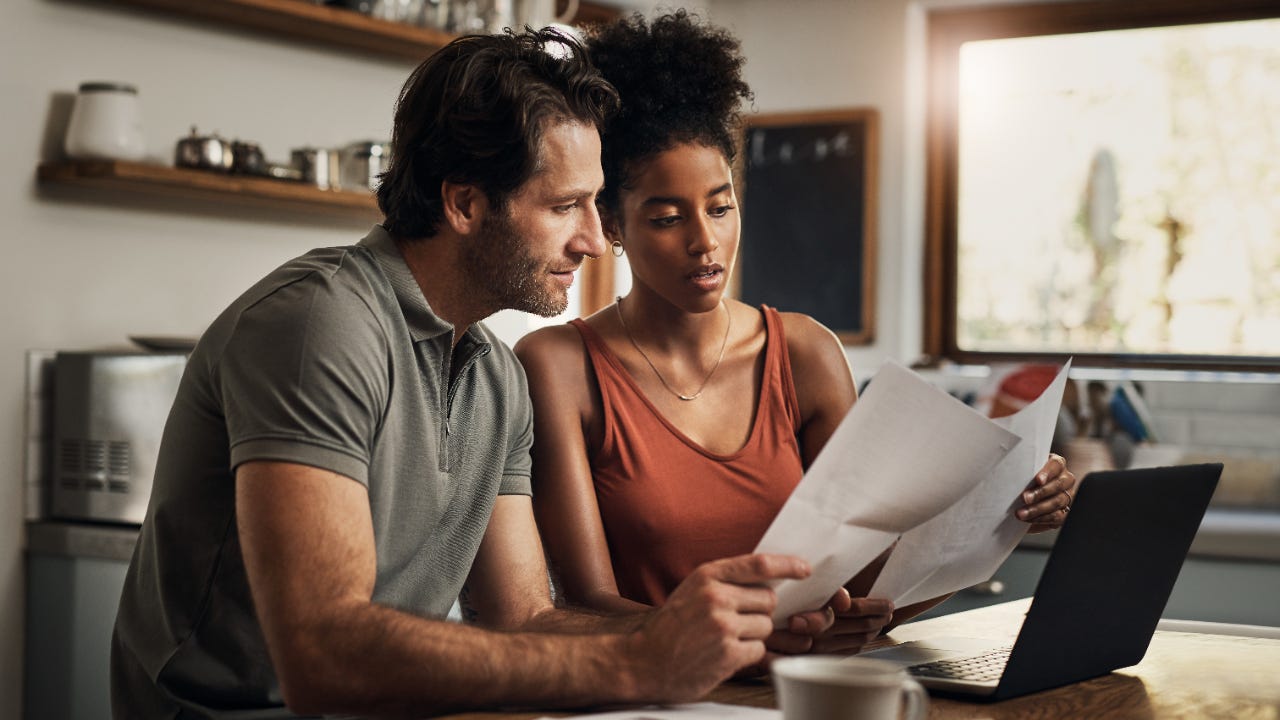 Couple sitting at a kitchen table review documents together, a laptop is open on the table.