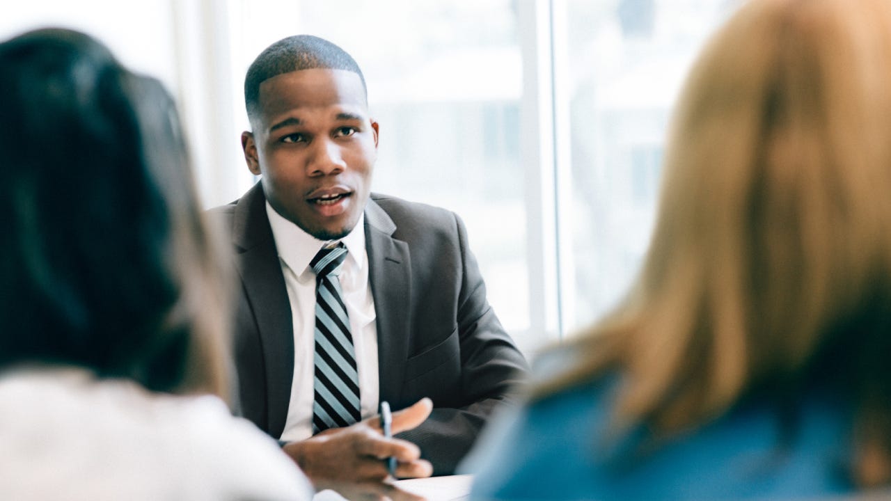 A black financial expert sits in his office with two clients, discussing some financial matters.