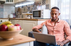 Man sitting on couch with laptop at home smiling, kitchen in background