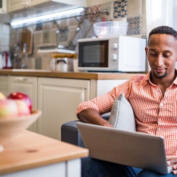 Man sitting on couch with laptop at home smiling, kitchen in background
