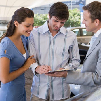 Young white man and woman standing with a car salesperson, signing a document