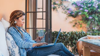 Woman sitting on patio with open laptop on lap and phone in hand