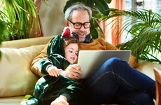 Father and son (who is dressed like a dinosaur) sit on the couch in front of a laptop looking at funny viral videos.
