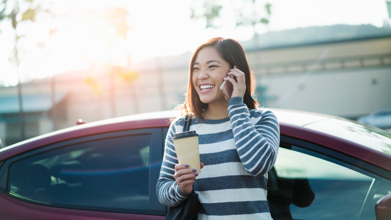 Young woman on phone leaning against car and holding coffee