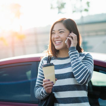 Young woman on phone leaning against car and holding coffee