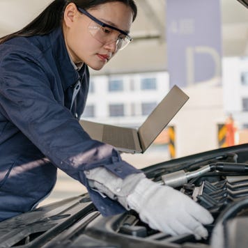 Woman in safety goggles holding a laptop in her left hand and checking under a car hood