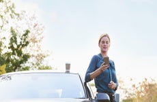 Woman leaning on her car looking at her phone with a coffee atop the car