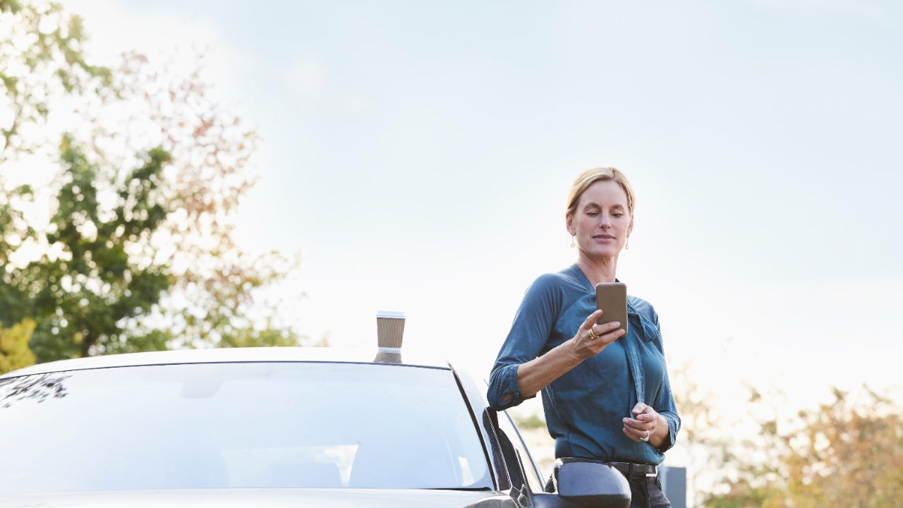 Woman leaning on her car looking at her phone with a coffee atop the car
