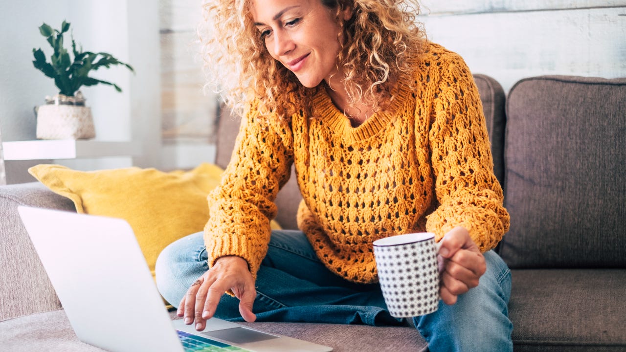 Smiling woman in yellow sweater sits on couch with coffee mug in hand while using laptop
