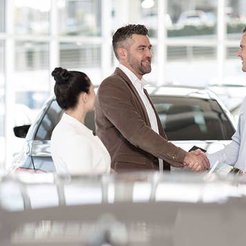 Man and woman at a car dealership talking to a salesperson. The man is shaking hands with the salesperson.