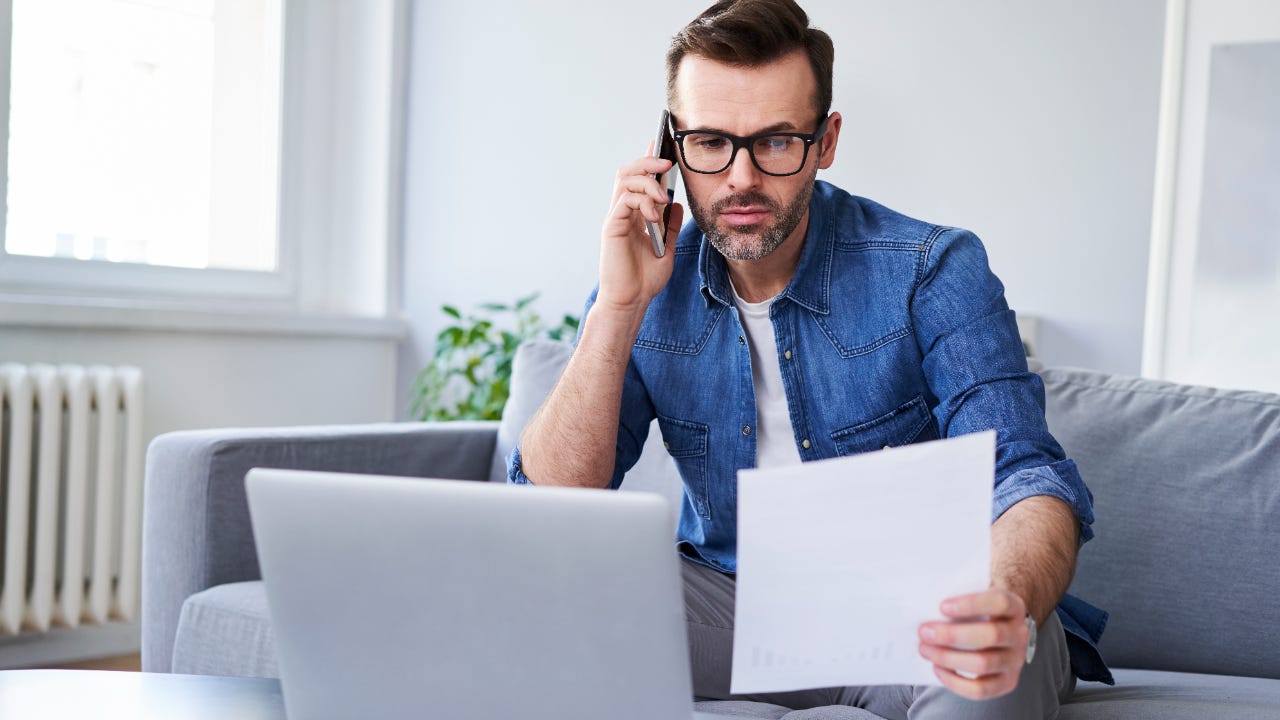 Serious man with glasses on phone looks at paper with laptop open in front of him