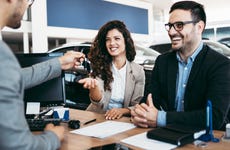 Man behind desk hands car key to smiling man and woman sitting on the other side with cars behind them