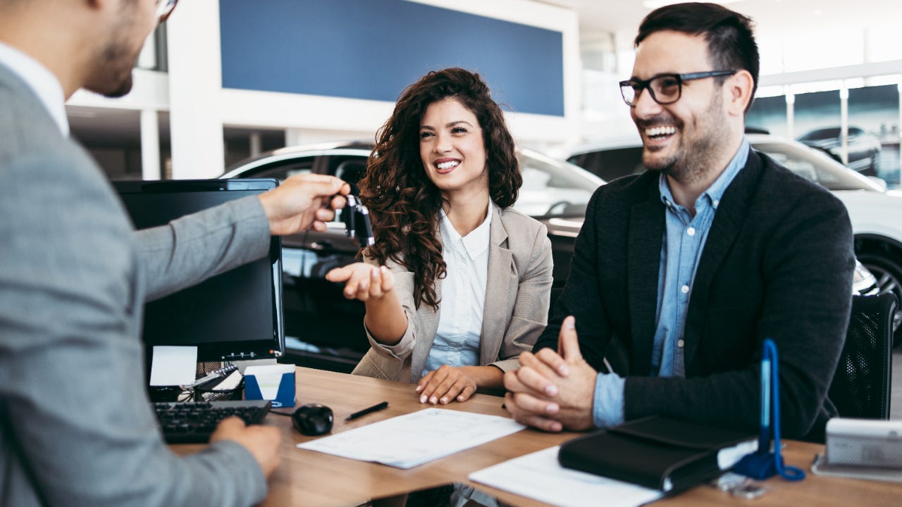 Man behind desk hands car key to smiling man and woman sitting on the other side with cars behind them