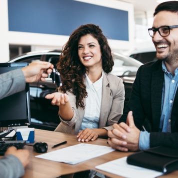 Man behind desk hands car key to smiling man and woman sitting on the other side with cars behind them