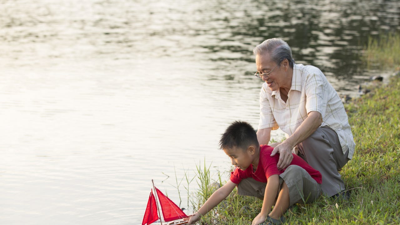 Asian grandfather and son hang out by the lake and put a toy sailboat into the water.
