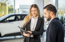 Woman showing man tablet while standing in front of car in dealership