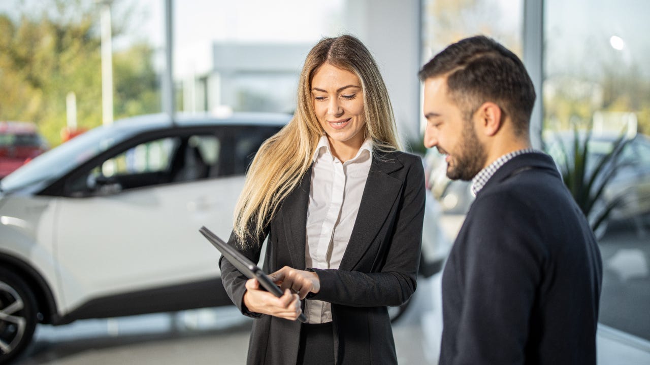 Woman showing man tablet while standing in front of car in dealership