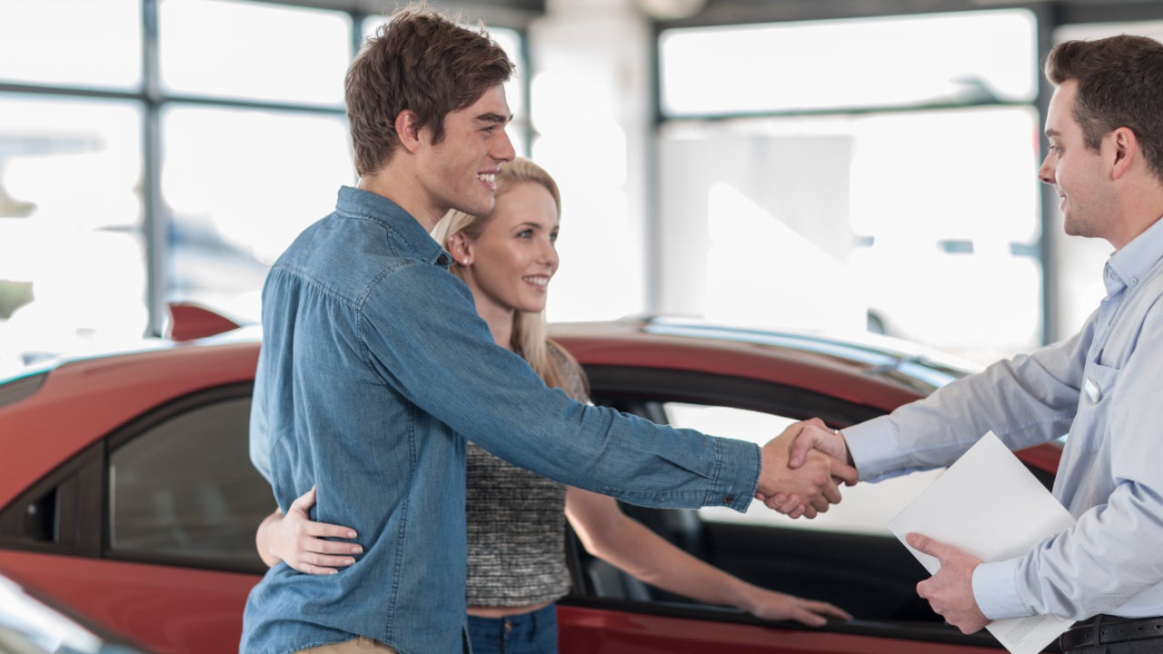 Woman standing next to car with arm around man who is shaking the hand of a car dealer