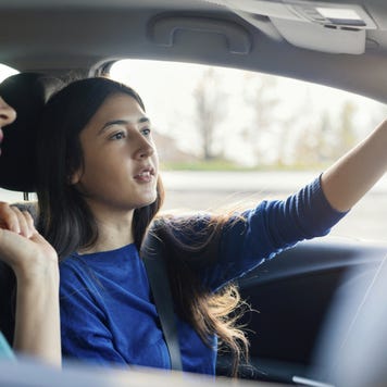 A teenage girl is adjusting the rearview mirror in her car while her mom is sitting in the passenger seat.