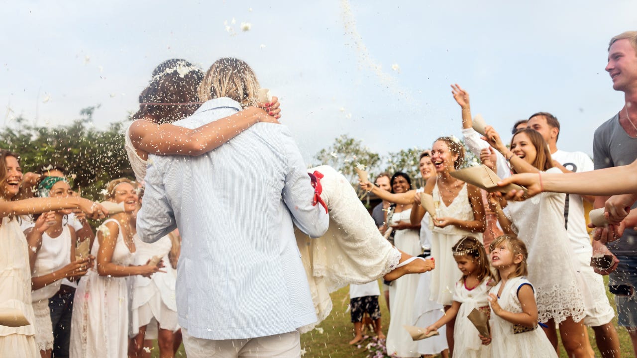 The back of a groom carrying his bride at an outdoor wedding while attendees throw rice!