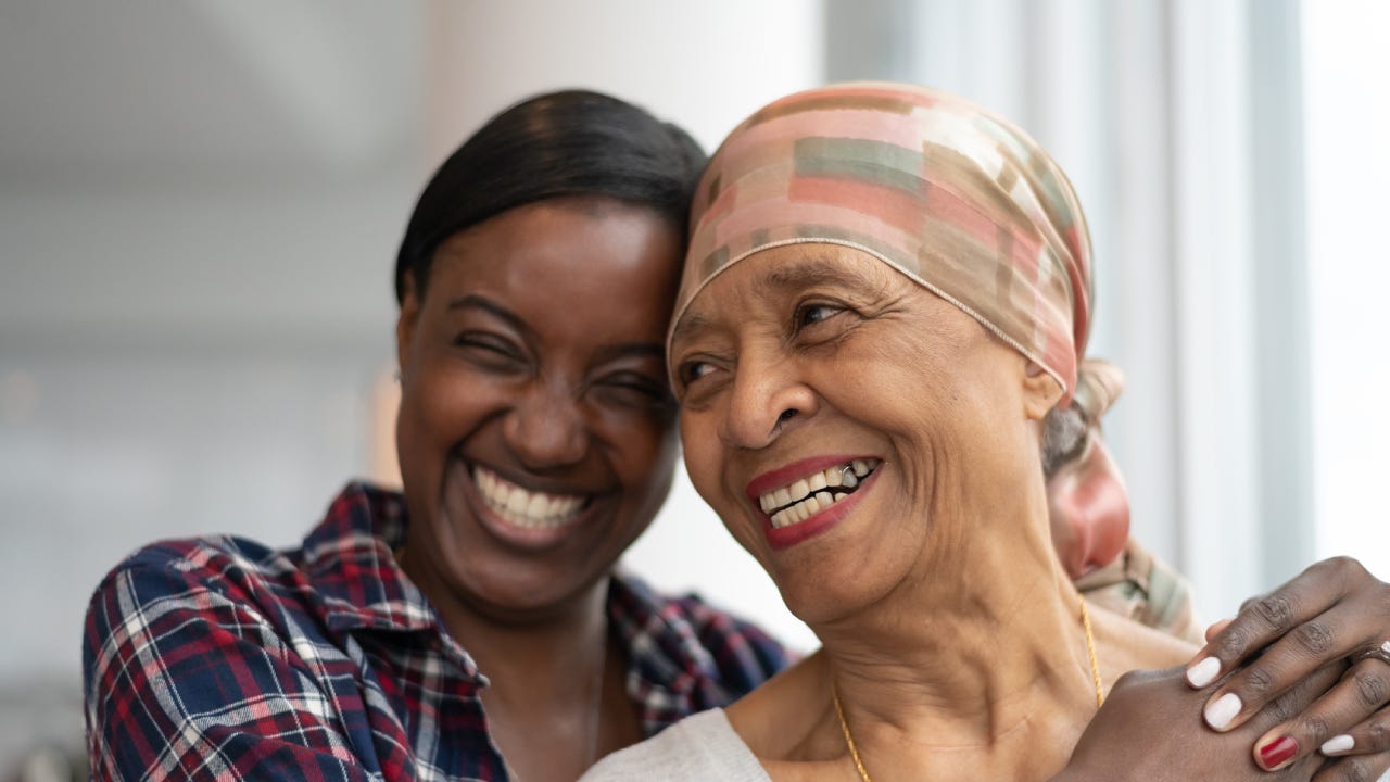 A mother and daughter smile together for a picture. The mother is wearing a silk scarf over hear head as she is undergoing chemotherapy.