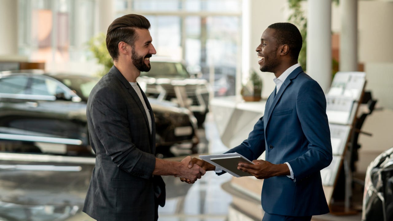 Two men in suits smiling and shaking hands in a car dealership