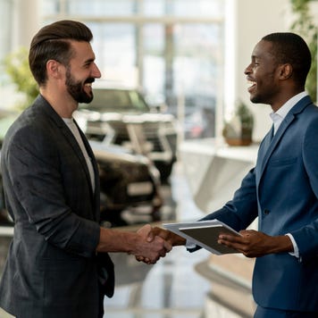 Two men in suits smiling and shaking hands in a car dealership