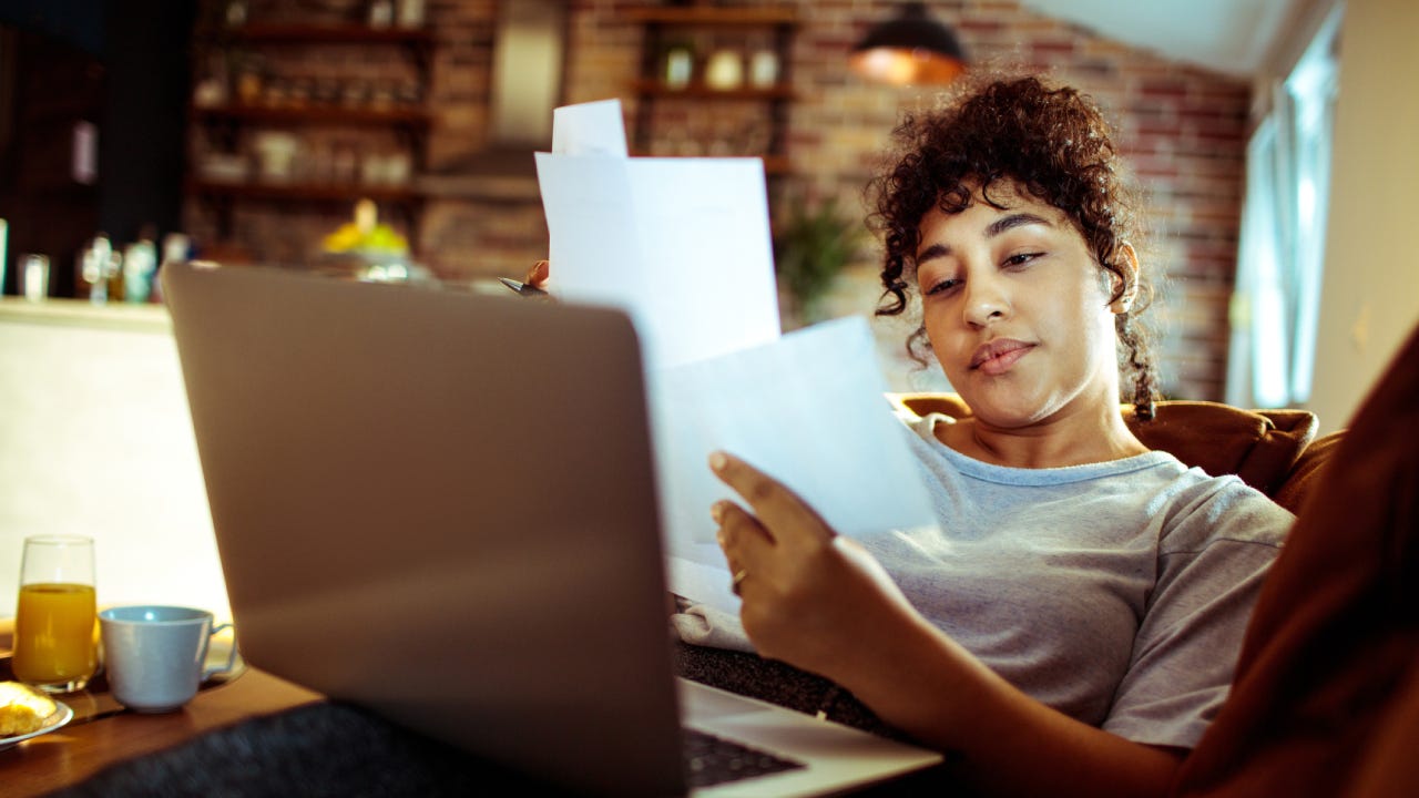 A woman lounges on her couch with a laptop on her lap and documents in her hands trying to make sense of it all.