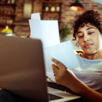 A woman lounges on her couch with a laptop on her lap and documents in her hands trying to make sense of it all.