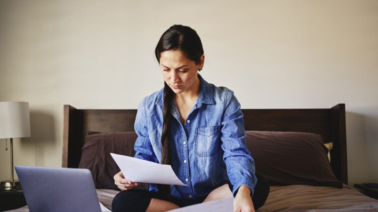 A young woman is sitting cross-legged on a couch and reviewing some financial forms.